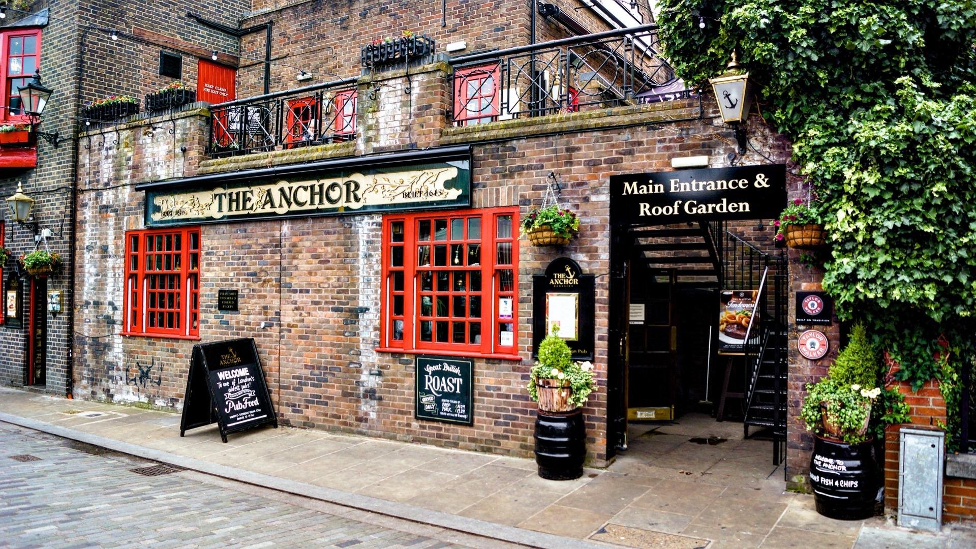 Exterior view of "The Anchor" pub, featuring a red and black brick facade with several windows displaying red frames. Above the main entrance, a sign reads "Main Entrance & Roof Garden". Other details include potted plants, hanging flower baskets, and a blackboard advertising food and drinks. The establishment has decorative greenery overhanging the entrance and a red balcony above.