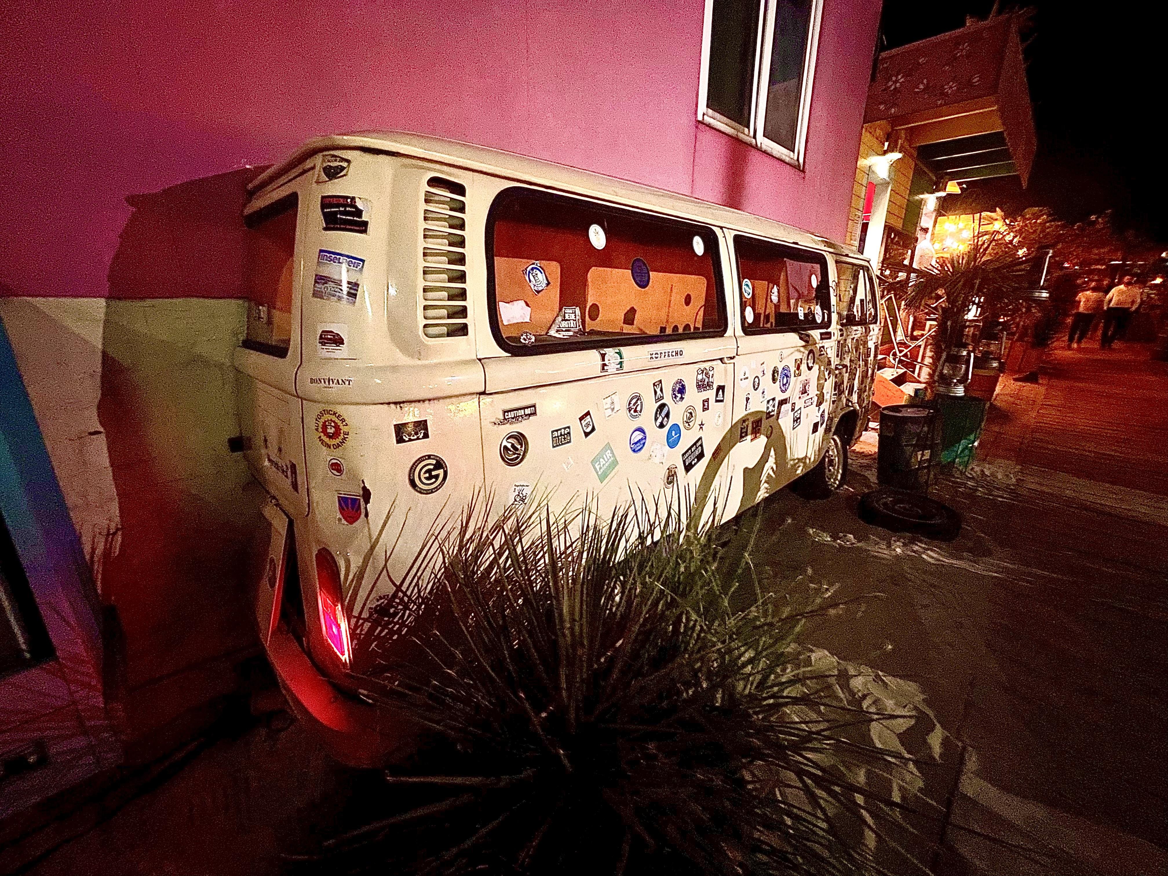 A vintage white Volkswagen van with multiple stickers on its side and rear windows is parked beside a pink wall with a green-painted curb. The van's brake light is illuminated red, casting a glow on the ground. In the foreground, there's a spiky plant, possibly an agave, and the background shows a night setting with low lighting and a few indistinct figures walking near colorful storefronts.