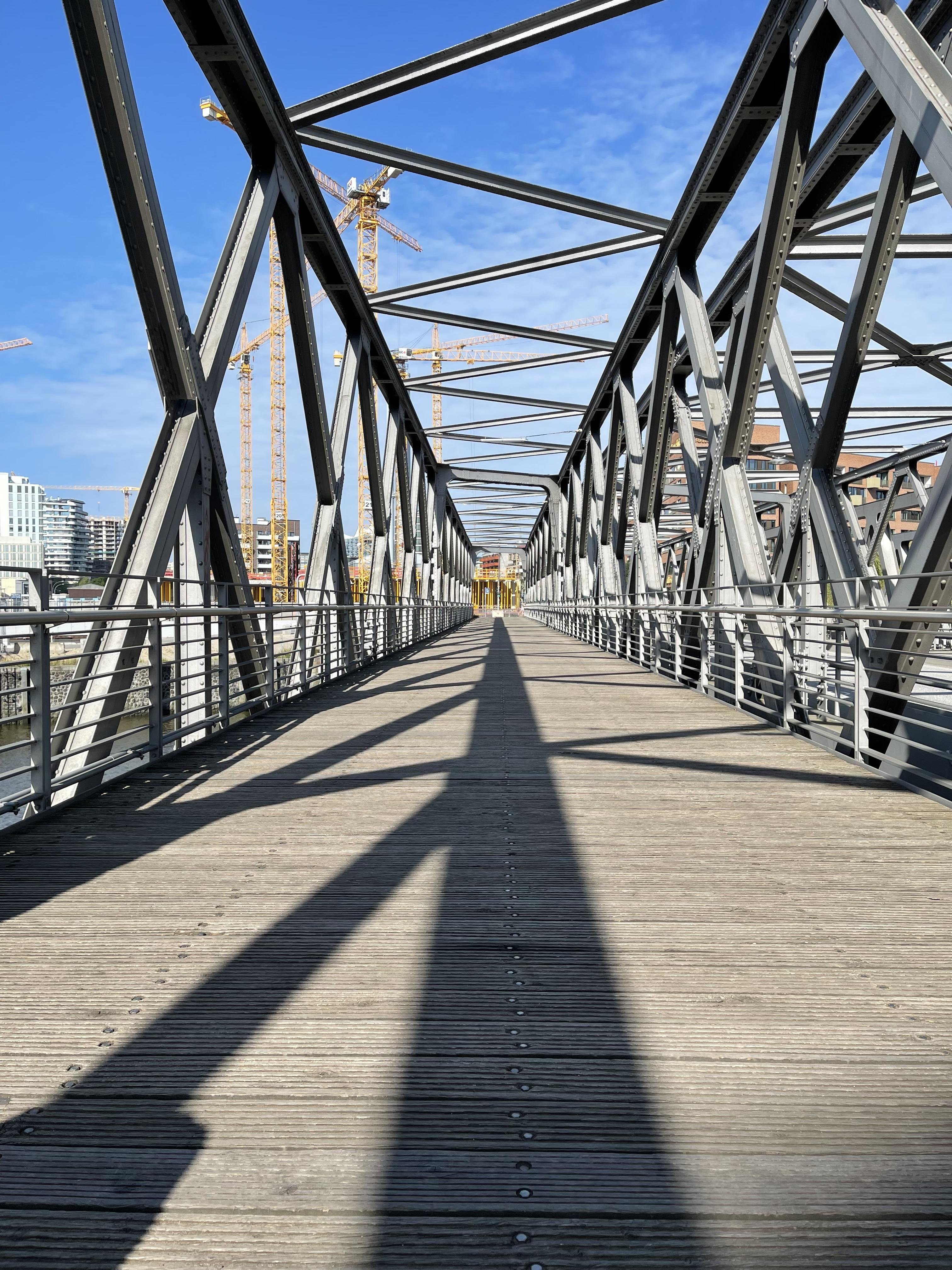 A metal truss bridge with a wooden walkway is depicted under a clear blue sky. The geometric pattern formed by the metal beams creates a converging perspective towards the center of the image, where construction cranes are visible in the background. Long shadows stretch across the walkway, emphasizing the bridge's linear design.
