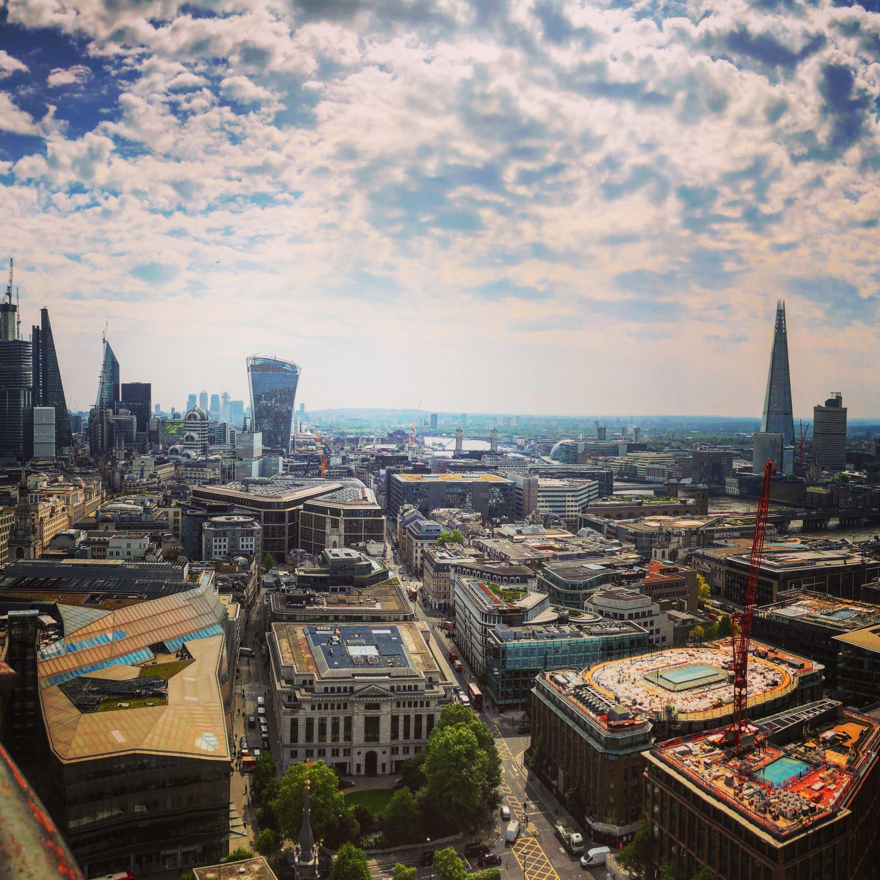 Aerial view of a cityscape on a clear day, featuring a mixture of modern skyscrapers and traditional buildings under a partly cloudy sky. Notable landmarks include a tall glass skyscraper resembling a shard, a distinctive building shaped like a walkie-talkie, and a large construction crane. The horizon shows distant views of a river and more urban development.